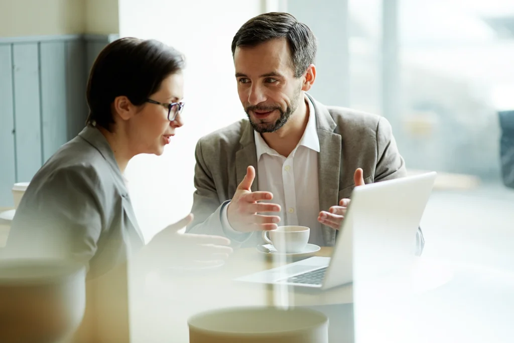 Zwei Geschäftsleute im Gespräch über Vertriebsstrategie am Laptop im Café.